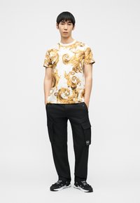 Νεαρός άνδρας στέκεται με τα χέρια στις τσέπες, φορώντας λευκό και χρυσό T-shirt με μπαρόκ σχέδιο, μαύρο παντελόνι cargo και μαύρα αθλητικά παπούτσια σε λευκό φόντο.