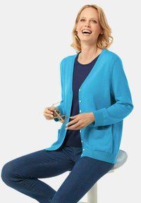 Türkiser Cardigan mit Knopfleiste, gerippten Bündchen und zwei vorderen Taschen, über einem marineblauen Oberteil getragen, kombiniert mit dunklen Jeans.