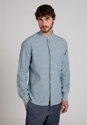 Mężczyzna stoi, mając na sobie jasnoniebieską koszulę z kołnierzykiem mandarynkowym oraz ciemnoniebieskie spodnie na jednolitym tle.