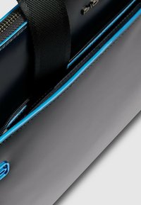 Bolso de cuero negro con acabado brillante, con un ribete de cremallera azul. Incluye una correa duradera de tela negra para llevar.