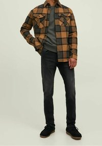 Camisa de franela a cuadros en marrón y negro, superpuesta sobre una camiseta de cuello redondo gris, combinada con jeans oscuros y zapatos negros.