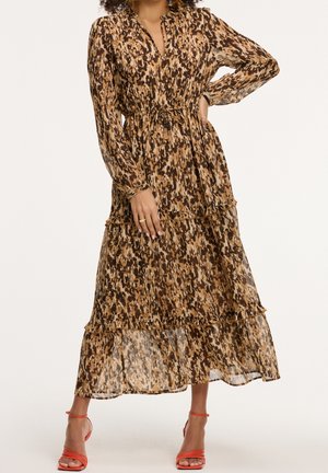Femme en robe longue à manches longues, marron à motifs, à volants avec ceinture à la taille, posant avec une main sur la hanche, portant des talons à brides orange.