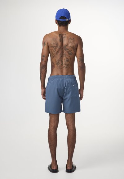 Shorts de bain bleu avec une taille élastique et une poche arrière, vus de dos. Modèle portant une casquette bleue, affichant des tatouages complexes.