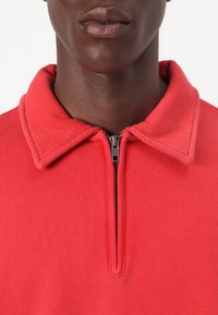 Maglietta rossa con zip, caratterizzata da un ampio colletto e una texture a coste. La cerniera è posizionata centralmente, in armonia con il colore e il design.