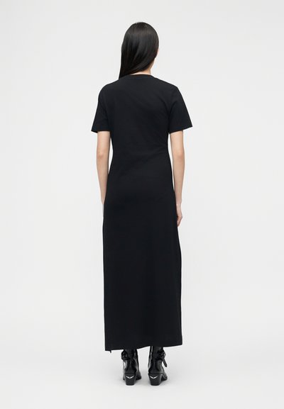 Γυναίκα με μακριά μαύρα μαλλιά, εμφανισμένη από πίσω, που φοράει μαύρο μαξι φόρεμα με κοντά μανίκια και μαύρα μποτάκια μέχρι τον αστράγαλο σε λευκό φόντο.