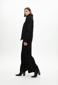 Černý pletený svetr s vysokým límcem a zlatými knoflíkovými detaily, spárovaný s širokými černými kalhotami se zapínáním na boku a černými lodičkami.