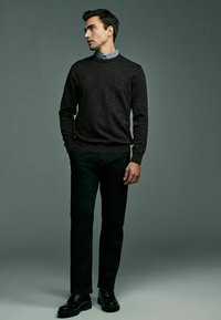 Pull en maille gris foncé sur une chemise col v bleu clair, associé à un pantalon noir et des chaussures noires, se tenant sur un fond neutre.