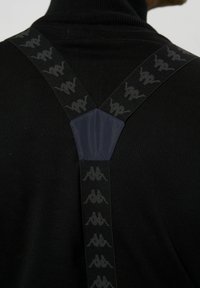 Črne naramnice z teksturirano tkanino, ki imajo vzorec logotipa in enobarven mornarsko moder osrednji del. Ozadje je temna rolka.