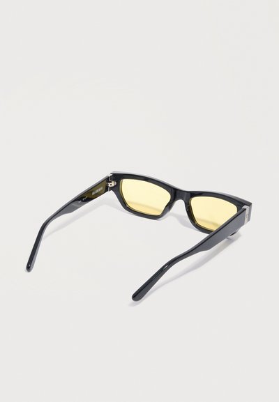 Μαύρα ορθογώνια γυαλιά ηλίου με λεπτά πλαίσια και φακούς με κίτρινη απόχρωση, εμφανιζόμενα από πίσω σε ανοιχτό φόντο.