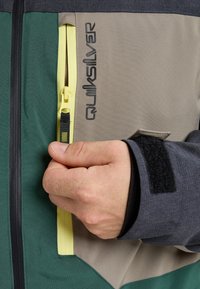 Hand, die den gelben Reißverschluss einer Quiksilver-Jacke in Grün, Beige und Grau hält, mit einem schwarzen Klettverschluss am Ärmel.