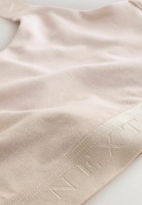 Ljusbeige tyg-bralette med en slät yta, som har ett brett band med präglad text längs nedre kanten.