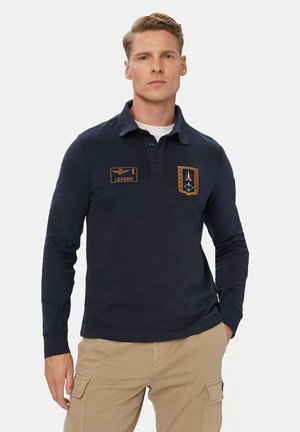 Uomo che indossa una polo blu navy a maniche lunghe con due toppe ricamate e pantaloni cargo beige, in piedi davanti a uno sfondo semplice.