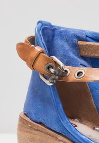 Detailní záběr modré semišové sandály s hnědým koženým páskem na kotníku a texturovanou kovovou přezkou na dřevěné podrážce na bílém pozadí.