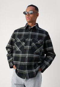 Svart- og hvitrutete skjorte med to brystlommer, knappelukking og en myk tekstur, båret med slanke lysegrå jeans og ovale solbriller.