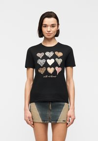 Νεαρή γυναίκα που φοράει μαύρο t-shirt με εννέα καρδιές σε σχέδιο ζώου και το κείμενο "αγριεμένη στην καρδιά", συνδυασμένο με δίχρωμη μίνι φούστα από τζιν με φερμουάρ μπροστά.