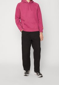 Roze hoodie met een kangoeroezak en een capuchon met trekkoord, gecombineerd met zwarte broek en sneakers met zwart-witte accenten.