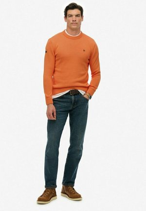 Hombre de pie con las manos en los bolsillos, vistiendo un suéter naranja, jeans azules, botas marrones y un reloj de pulsera sobre un fondo blanco.
