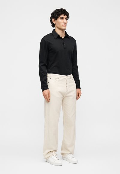 Άνδρας όρθιος, φορώντας μαύρο μακρυμάνικο πουκάμισο με γιακά, άνετο μπεζ παντελόνι και λευκά αθλητικά παπούτσια, μπροστά σε απλό φωτεινό φόντο.