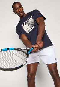 Ναυτικό t-shirt με γραφική εκτύπωση, λευκό σορτσάκι, κρατώντας ένα ρακέτα τένις με μπλε λεπτομέρειες. Απαλή υφή, σχεδίαση casual αθλητικών ρούχων.