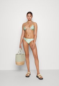 Conjunto de biquíni com padrão floral verde, top triangular e parte de baixo com cintura baixa. A modelo segura uma bolsa de palha trançada e usa sandálias pretas com detalhe de babado.