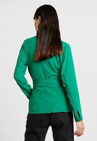 Grüne Bluse mit langen Ärmeln, tailliertem Schnitt und offenen Bündchen. Glattes Material, minimalistisches Design, keine sichtbaren Muster und eine elegante Silhouette.