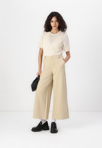 Beige wijde broek met hoge taille en een knoopdetail, gecombineerd met een crèmekleurig gebreid T-shirt met korte mouwen en zwarte instappers.