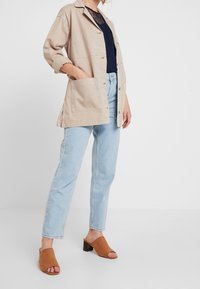 Veste en coton beige à coupe décontractée, avec de grandes poches avant et une fermeture à boutons. Associée à un jean droit bleu clair et des mules marron.