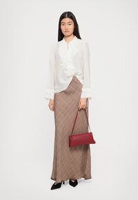 Hvit, volangbluse kombinert med et brunt mønstrete maxiskjørt. Modellen holder en rød clutch-veske og har på seg svarte hæler. Jevn tekstur og strukturert design.
