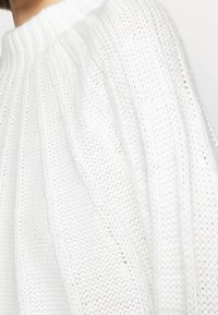 Pull en tricot blanc avec un design côtelé et un col montant texturé. Le tissu semble doux et léger avec une coupe ample.