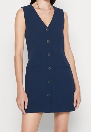 Femme portant une robe sans manches bleu marine à boutons, avec un col en V et deux poches devant, debout devant un fond clair uni.