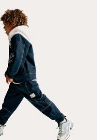 Marineblauwe hoodie met witte accenten en joggingsbroek, met contrasterende stiksels en een logo-label, gecombineerd met grijze en witte sneakers.