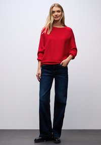 Rode geribbelde trui met driekwart mouwen, gecombineerd met donkerblauwe wijde jeans. Model staat casual met handen in de zakken op een effen achtergrond.