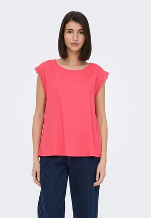 Donna con capelli lisci e scuri indossa un top rosa corallo senza maniche con rifiniture in pizzo e jeans blu scuro, in piedi contro uno sfondo chiaro.
