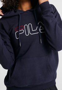 Felpa blu navy con tasca frontale, cordini e logo "FILA" bianco e rosso sul petto. Tessuto morbido.
