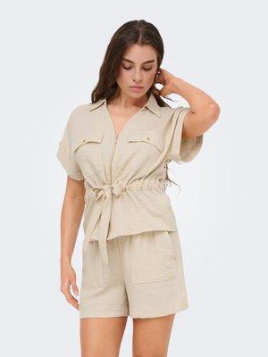 Vrouw met lang bruin haar, gekleed in een beige shirt met korte mouwen en rits aan de voorkant, een ceintuur in de taille en bijpassende shorts, die met één hand haar haar aanraakt.
