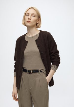 Femme aux cheveux blonds courts portant un cardigan marron, une chemise taupe à manches longues et un pantalon plissé beige avec ceinture noire, debout, une main dans la poche.