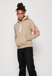 Beige hettegenser med frontlomme, logo på brystet, og skulderlomme med rød glidelås. Sorte bukser fullfører antrekket.