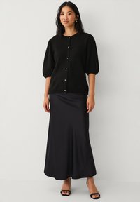 Cardigan nero lavorato a maglia con maniche a sbuffo corte, dotato di bottoni in perla. Abbinato a una gonna maxi nera liscia e sandali con tacco alto.