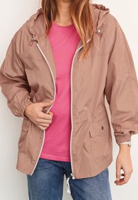 Lightweight, hooded jacket in dusty rose, featuring a zip front, elastic cuffs, and side pockets. Worn over a pink shirt and jeans.