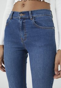 Vaqueros de mezclilla azul con un diseño ajustado, que cuentan con costuras naranjas, cinco bolsillos y un cierre de botón en la cintura. Textura suave.