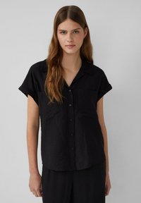 Schwarzes kurzärmeliges Hemd mit Knöpfen, das einen Hemdkragen, zwei vordere Taschen und eine strukturierte Stoffoberfläche aufweist.