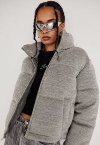 Ung person med fletninger bærer sølvsolbriller, guld hoopsøreringe, næse-ringe, en grå pufferjakke, sort top og udslidte jeans.