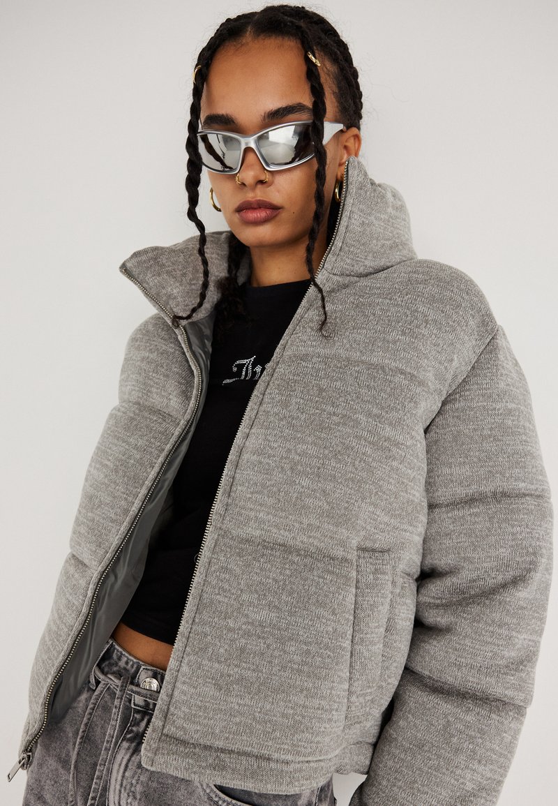 Ung person med fletninger bærer sølvsolbriller, guld hoopsøreringe, næse-ringe, en grå pufferjakke, sort top og udslidte jeans.