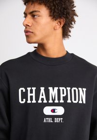 Hanorac negru cu text mare alb "CHAMPION" și "ATHL. DEPT." cu accent logo. Guler și manșete ribbate, textură netedă.