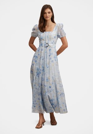 Žena v opásaných svetlomodrých maxi šatách s kvetinovým vzorom, nafúknutými rukávmi a štvorcovým výstrihom, stojaca s rukami za chrbtom.