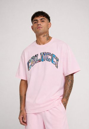 Man met tatoeages die een roze t-shirt en shortset draagt, het t-shirt heeft kleurrijke "COLUCCI" tekst over de borst, staand tegen een effen achtergrond.