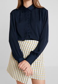 Camisa de manga larga color azul marino con botones en la parte delantera y cuello, combinada con una falda de cuadro amarilla y azul.