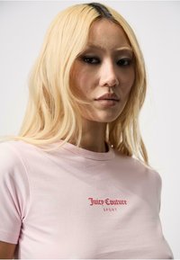 Gaiši rozā kokvilnas T-kreklis ar apaļu kakla izgriezumu un īsām piedurknēm; uz krūšu daļas ir sarkans izšuvums ar "Juicy Couture SPORT" logo.