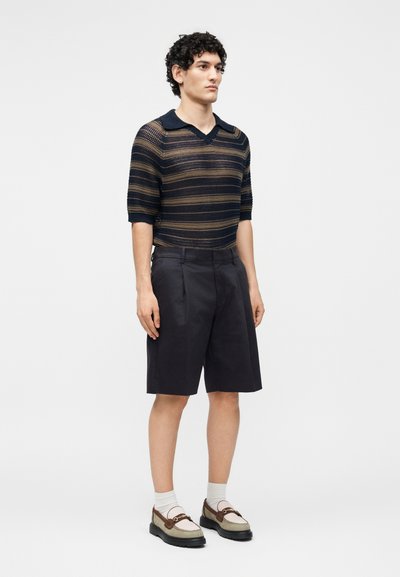 Homme portant un pull tricoté à manches courtes rayé, un short noir au genou, des chaussettes blanches et des mocassins bicolores, debout devant un fond uni.