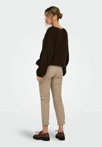 Blondiinide hobusesabaga naine, kellel on kuldsed rõngaskõrvarõnad, tumepruun lahtine kampsun, beežid mansetiga püksid ja pruunid nahast kingad.